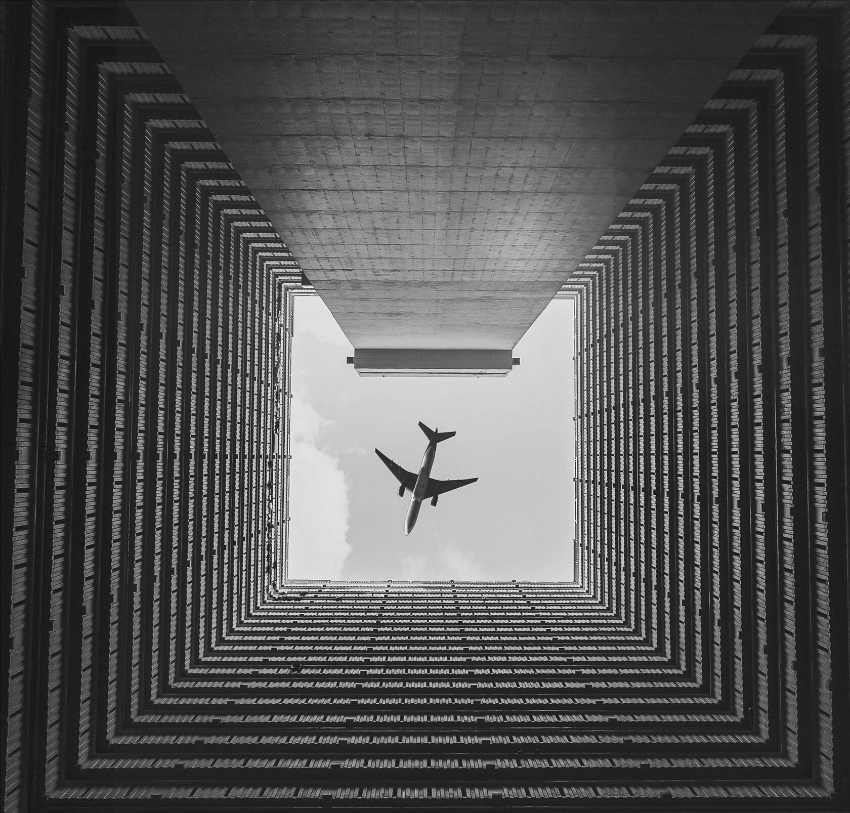 Чёрно-белая фотография, снятая снизу вверх, между высокими зданиями, образующими прямоугольный колодец. В центре неба пролетает самолёт, силуэт которого выделяется на фоне светлого пространства. Геометрическая симметрия зданий и динамика самолёта создают ощущение масштаба и движения. 