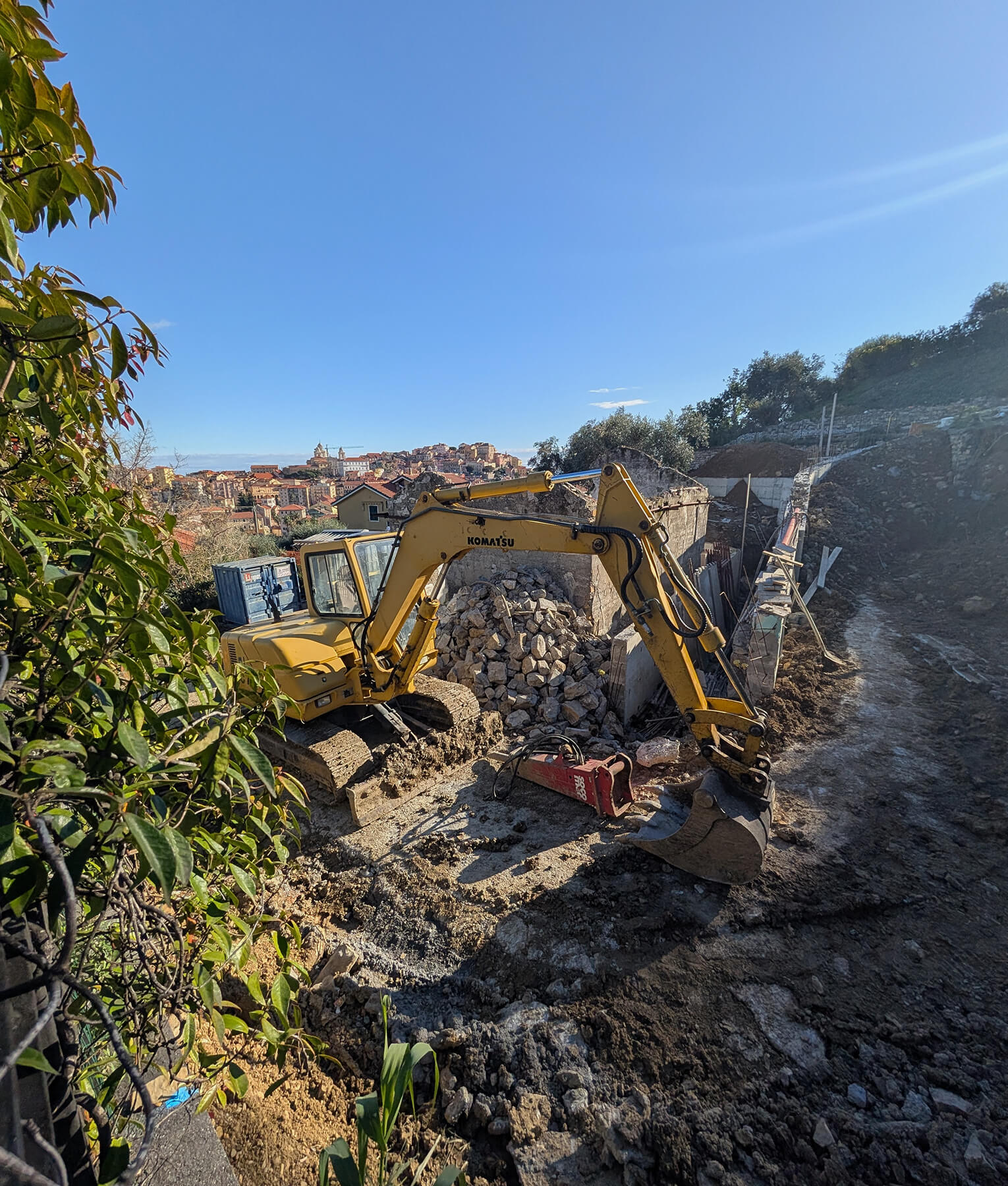 Excavator on hillside construction site in Liguria with mountainous landscape, and below, two men reviewing architectural plans and blueprints for renovation project supervision and management.