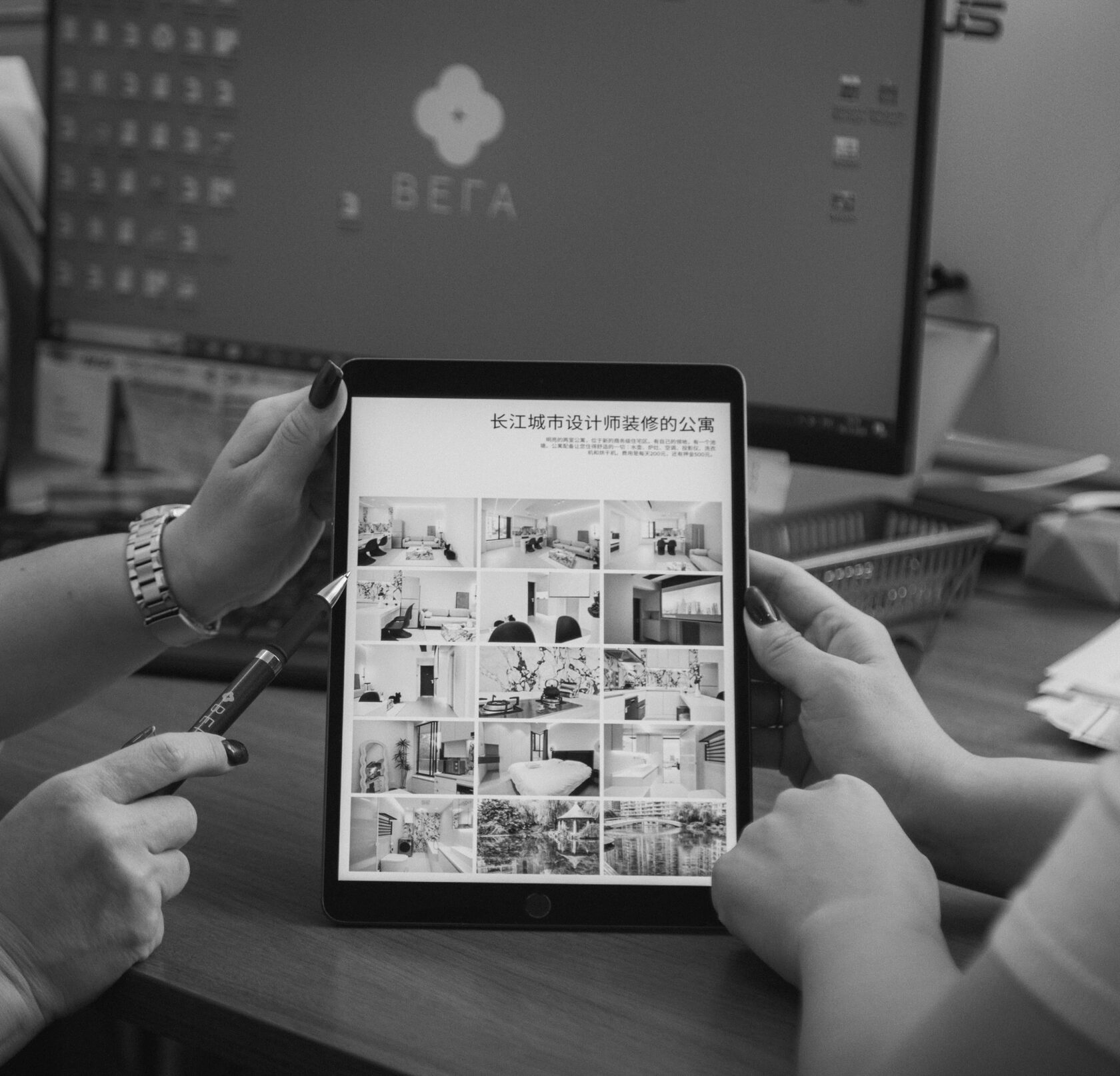 Чёрно-белая фотография двух человек, держащих планшет с изображениями интерьеров и текстом на китайском языке. На заднем плане виден монитор с логотипом компании "ВЕГА". Одна из рук указывает на планшет ручкой, создавая акцент на обсуждении или демонстрации контента. Снимок передаёт рабочую атмосферу и взаимодействие. 